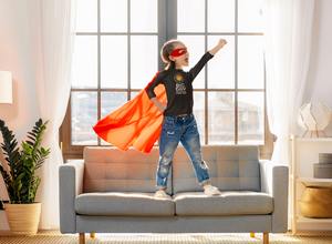 BIG DAY® Kids Apparel & Accessories Collection - girl wearing cape and mask standing on couch with fist in the air wearing BIG DAY Elevate Your Day black long sleeve.